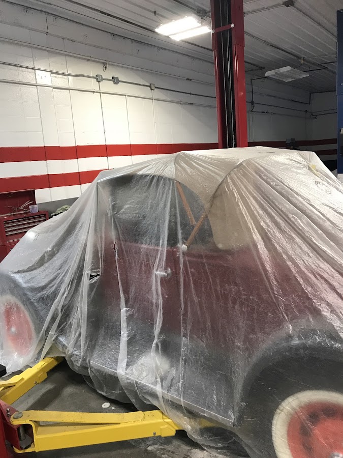 A car under protective plastic mid-paint job inside the shop, red and white striped walls visible